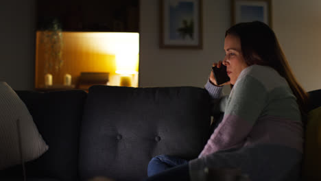 Smiling-Woman-Sitting-On-Sofa-At-Home-At-Night-Talking-On-Mobile-Phone-And-Watching-Movie-Or-Show-On-Laptop-3